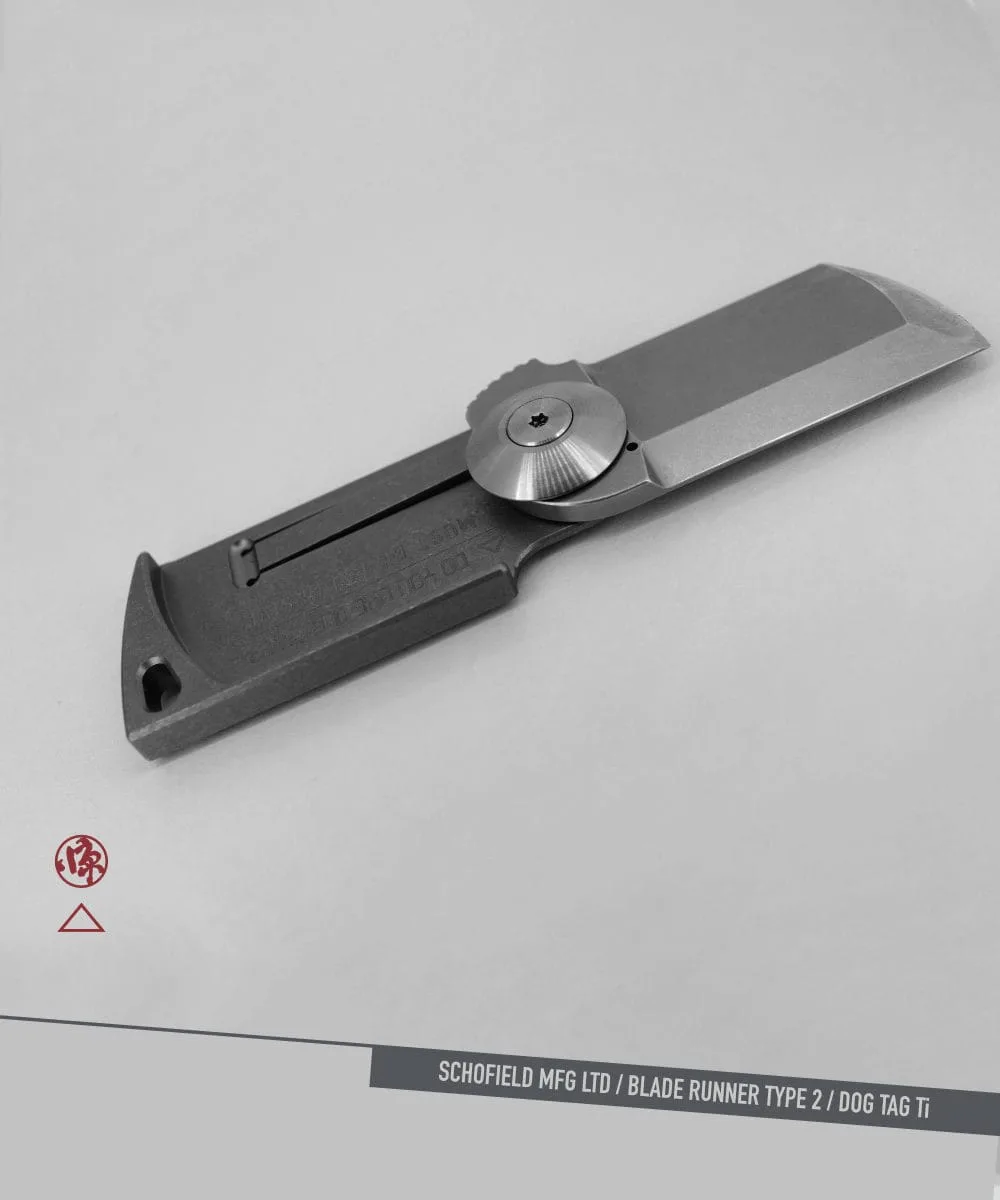 A compact, gray folding knife with a sleek, minimalist design is displayed on a light gray surface. Text reads "SCHOFIELD MFG LTD / BLADE RUNNER TYPE 2 / DOG TAG Ti" in the bottom right corner.