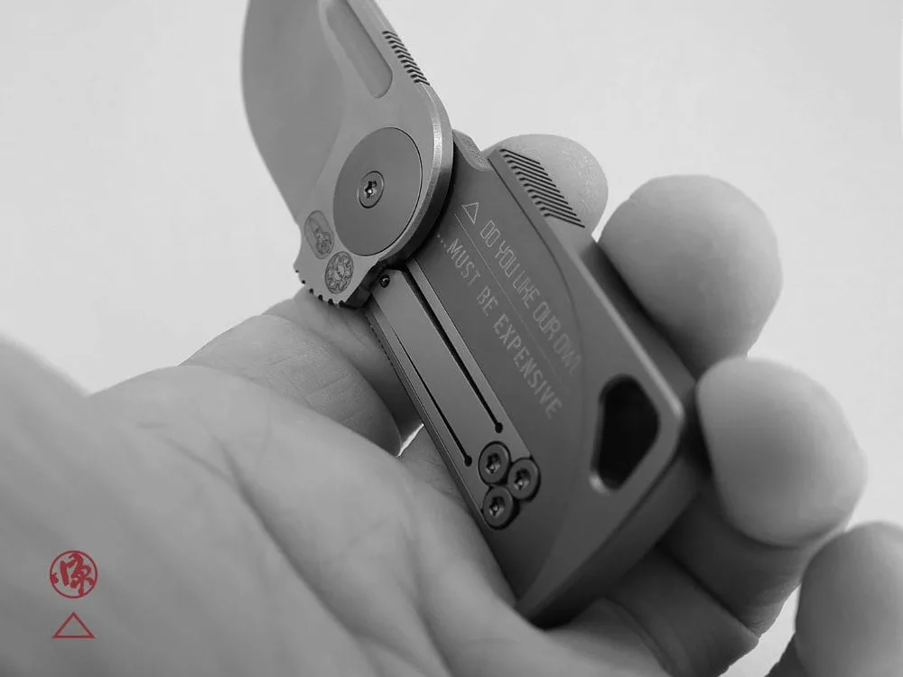 A hand holds a compact, folding knife with a short blade engraved "...MUST BE EXPENSIVE." The design is modern and made from titanium, with visible screws and a lanyard hole.