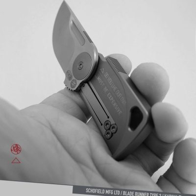 A hand holds a compact, folding knife with a short blade engraved "...MUST BE EXPENSIVE." The design is modern and made from titanium, with visible screws and a lanyard hole.