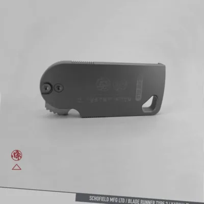 A compact, foldable utility knife with a gray metal body lies on a light surface. The words "SCHOFIELD MFG LTD / BLADE RUNNER TYPE 3 / KADIMA Ti" are printed along the bottom edge of the image.