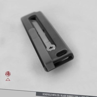 A black metal object with a rectangular shape and a visible screw is centered on a plain, light background. Text at the bottom reads: "SCHOFIELD MFG LTD / BLADE RUNNER TYPE 1 / ZIRC OTF.