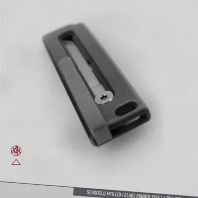 A black metal object with a rectangular shape and a visible screw is centered on a plain, light background. Text at the bottom reads: "SCHOFIELD MFG LTD / BLADE RUNNER TYPE 1 / ZIRC OTF.