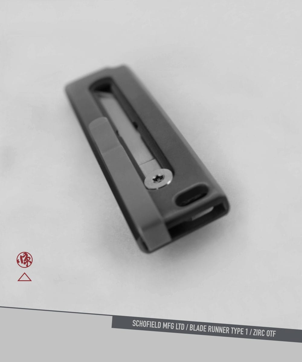 A black metal object with a rectangular shape and a visible screw is centered on a plain, light background. Text at the bottom reads: "SCHOFIELD MFG LTD / BLADE RUNNER TYPE 1 / ZIRC OTF. A black metal object with a rectangular shape and a visible screw is centered on a plain, light background. Text at the bottom reads: "SCHOFIELD MFG LTD / BLADE RUNNER TYPE 1 / ZIRC OTF.
