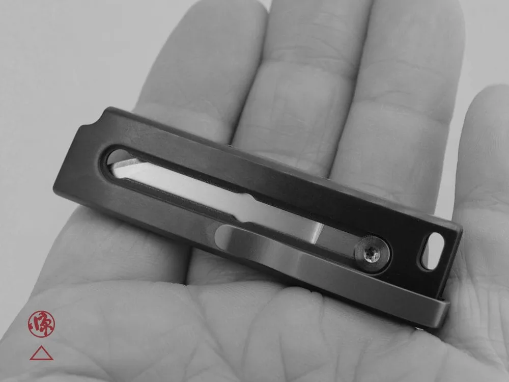 A person holds a compact, rectangular metal object with a slot and screw in the center. The background is a close-up of the person’s palm. Text at the bottom reads “SCHOFIELD MFG LTD / BLADE RUNNER TYPE 1 / ZIRC OTF.”.