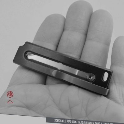 A person holds a compact, rectangular metal object with a slot and screw in the center. The background is a close-up of the person’s palm. Text at the bottom reads “SCHOFIELD MFG LTD / BLADE RUNNER TYPE 1 / ZIRC OTF.”.
