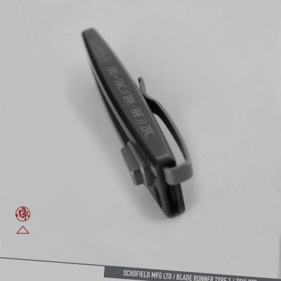 A dark metallic clip with engraved text rests on a light gray surface. Nearby are a small red logo and a label reading "SCHOFIELD MFG LTD / BLADE RUNNER TYPE 1 / ZIRC OTF.