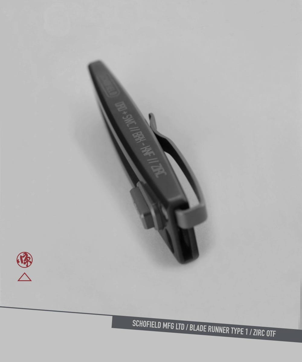 A dark metallic clip with engraved text rests on a light gray surface. Nearby are a small red logo and a label reading "SCHOFIELD MFG LTD / BLADE RUNNER TYPE 1 / ZIRC OTF. A dark metallic clip with engraved text rests on a light gray surface. Nearby are a small red logo and a label reading "SCHOFIELD MFG LTD / BLADE RUNNER TYPE 1 / ZIRC OTF.