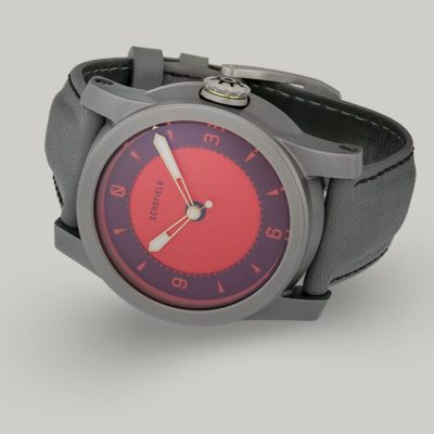 A grey titanium wristwatch with a red dial from Schofield. The Strange Lights ✦ NQR features custom lume on the Not Quite Red dial. Shown here on a grey strap with black stitching.