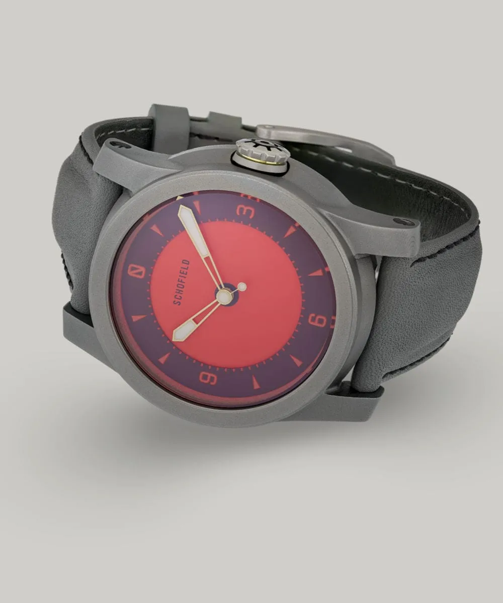 A grey titanium wristwatch with a red dial from Schofield. The Strange Lights ✦ NQR features custom lume on the Not Quite Red dial. Shown here on a grey strap with black stitching.